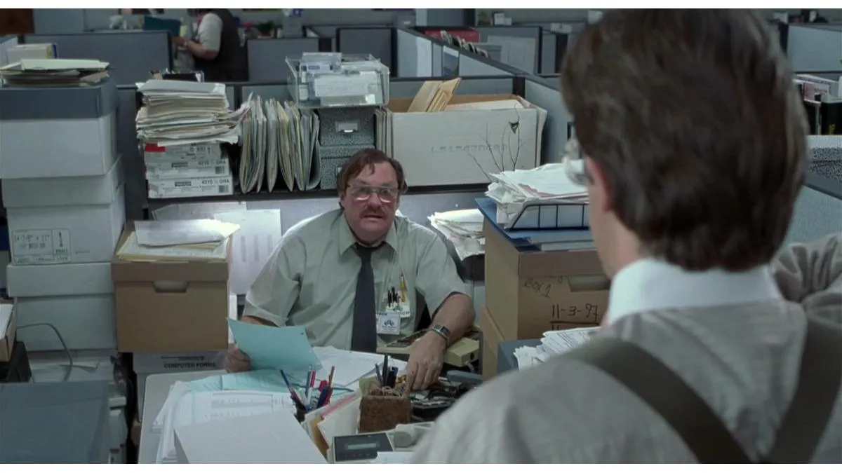 An office worker at a desk in a cluttered cubicle, surrounded by stacks of paper files and boxes